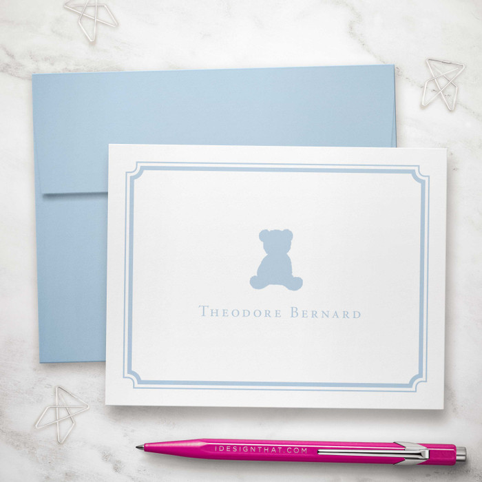 Personalized folded notecard featuring a soft blue teddy bear design with matching blue envelope. Shown with a pink pen and silver paper clips on a marble surface.